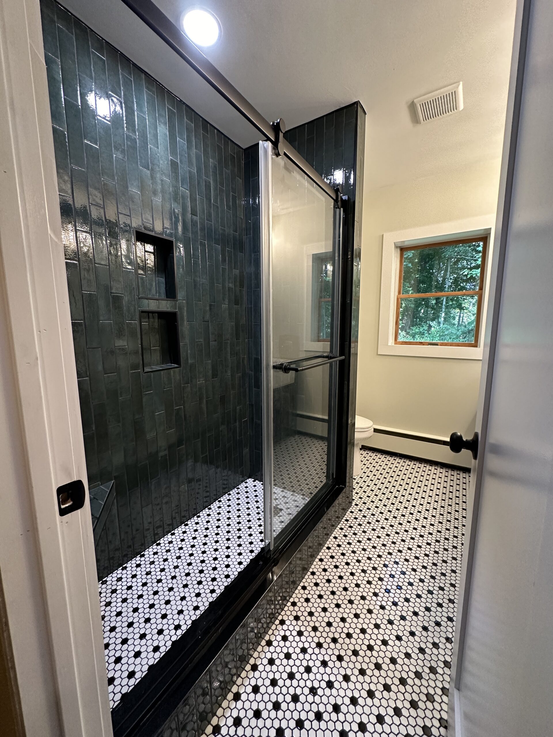 A modern bathroom with black tiled shower, glass doors, white walls, hexagon-patterned black and white floor tiles, and a window showing greenery outside.