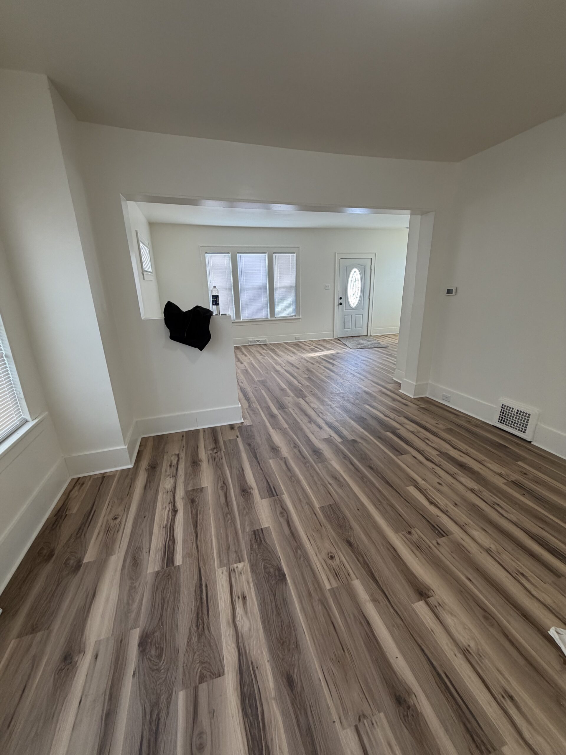 A view of an empty room with light walls and wood-patterned vinyl flooring, leading to another area with large windows and a white front door.